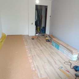 Yuniks - Układanie paneli podłogowych w jasnym wnętrzu. Część podłogi wyłożona korkiem, obok narzędzia i paczki paneli Aqua Block 24h. Widoczne białe ściany i sufit.