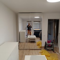 Yuniks - Nowoczesna szafa z lustrzanymi drzwiami przesuwnymi w jasnym pokoju. Widoczne białe komody i drabina malarska, sugerujące świeżo zakończone prace wykończeniowe.