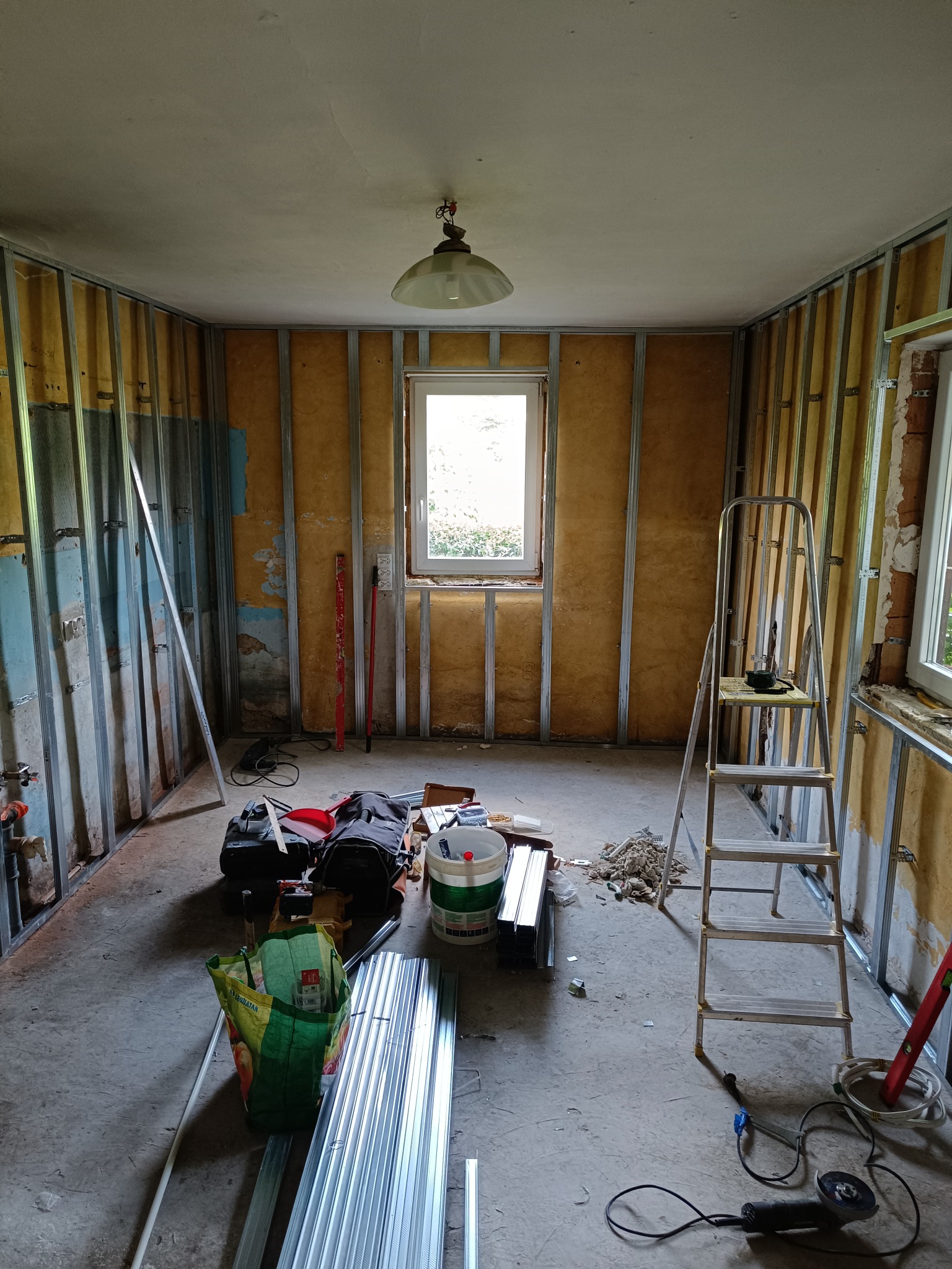 Wnętrze w trakcie montażu ścian działowych z profili stalowych. Widoczne narzędzia, drabina i materiały budowlane. Okno z widokiem na zieleń w tle.