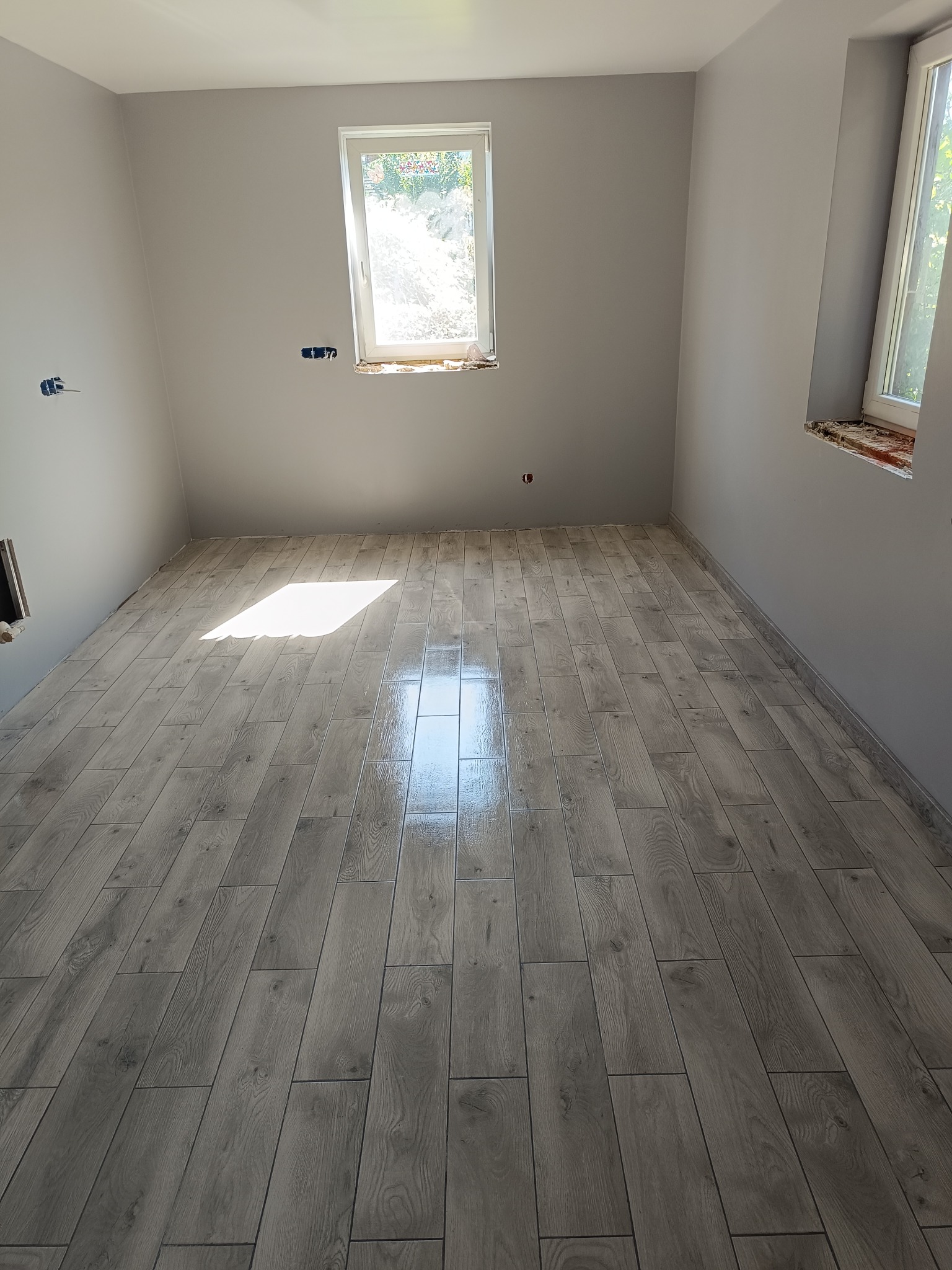 Wykończone wnętrze: szare ściany, drewnopodobne płytki podłogowe ułożone w jodełkę, dwa okna z widokiem na zieleń. Widoczne gniazdka elektryczne.