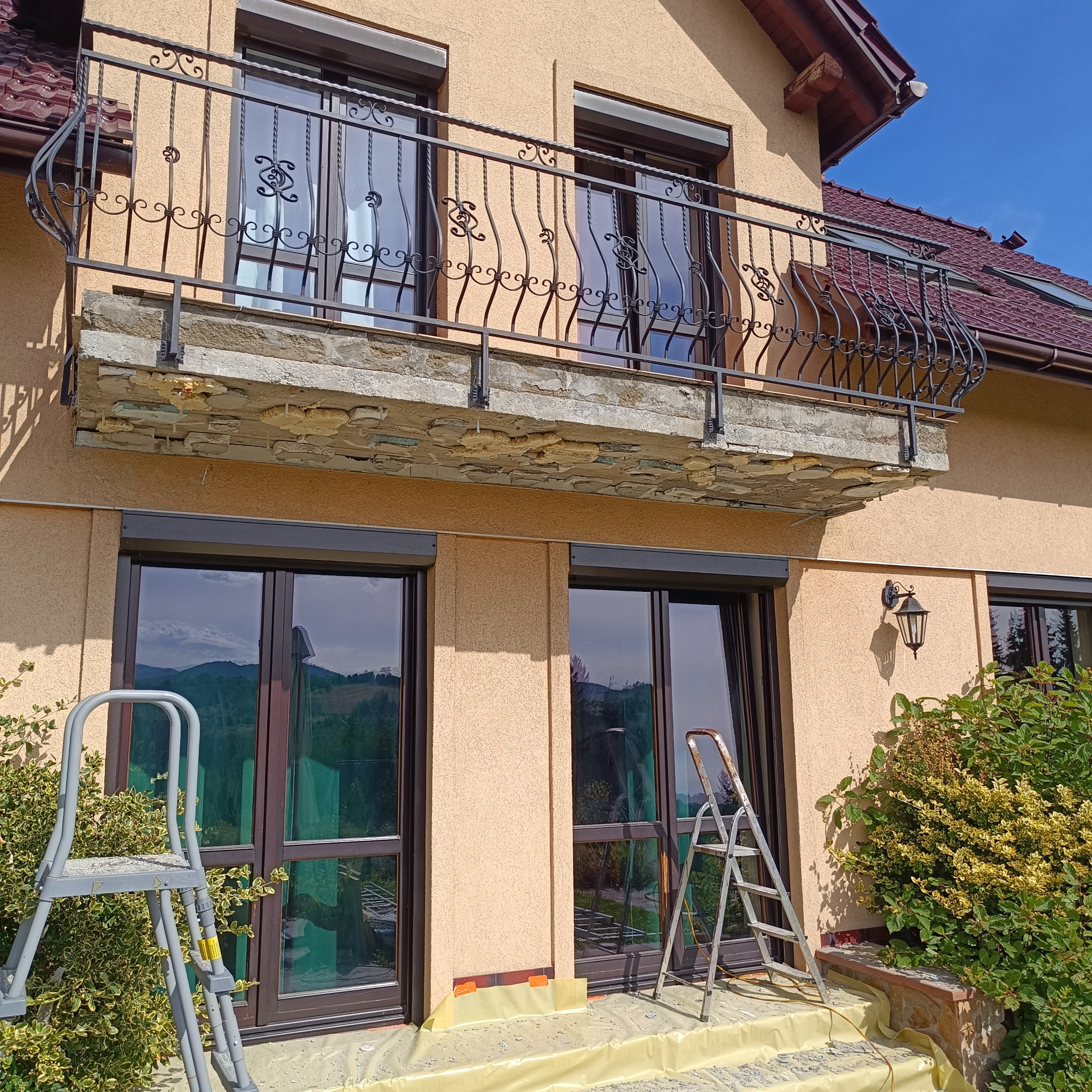 Elewacja domu z balkonem z ozdobną, kutą balustradą. Widoczne drabiny i folia ochronna sugerują prace remontowe. Okna z roletami w kolorze balustrady.