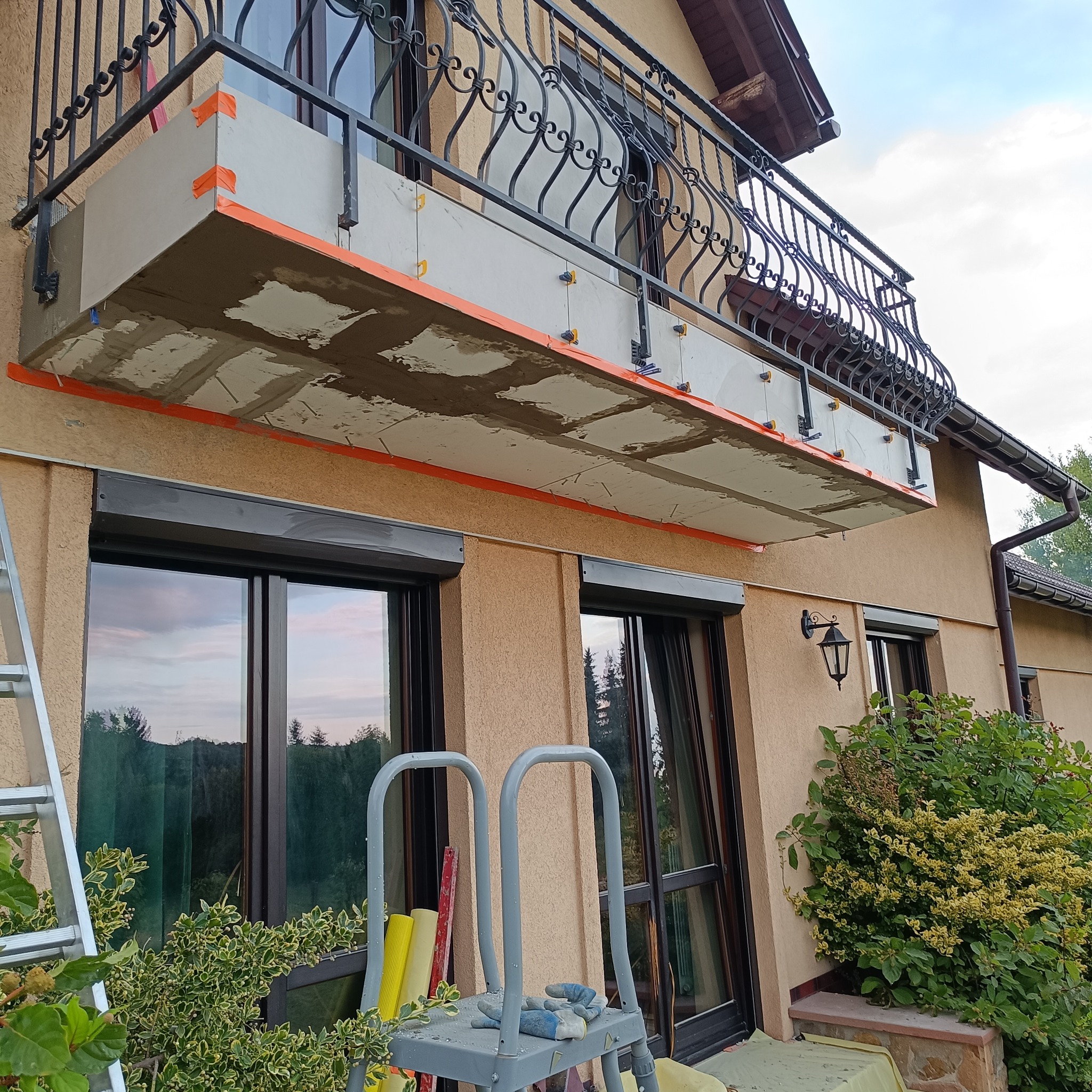 Balkon w trakcie prac wykończeniowych: płyty mocowane do sufitu, pomarańczowa taśma, rusztowanie, widok z dołu na elewację budynku.