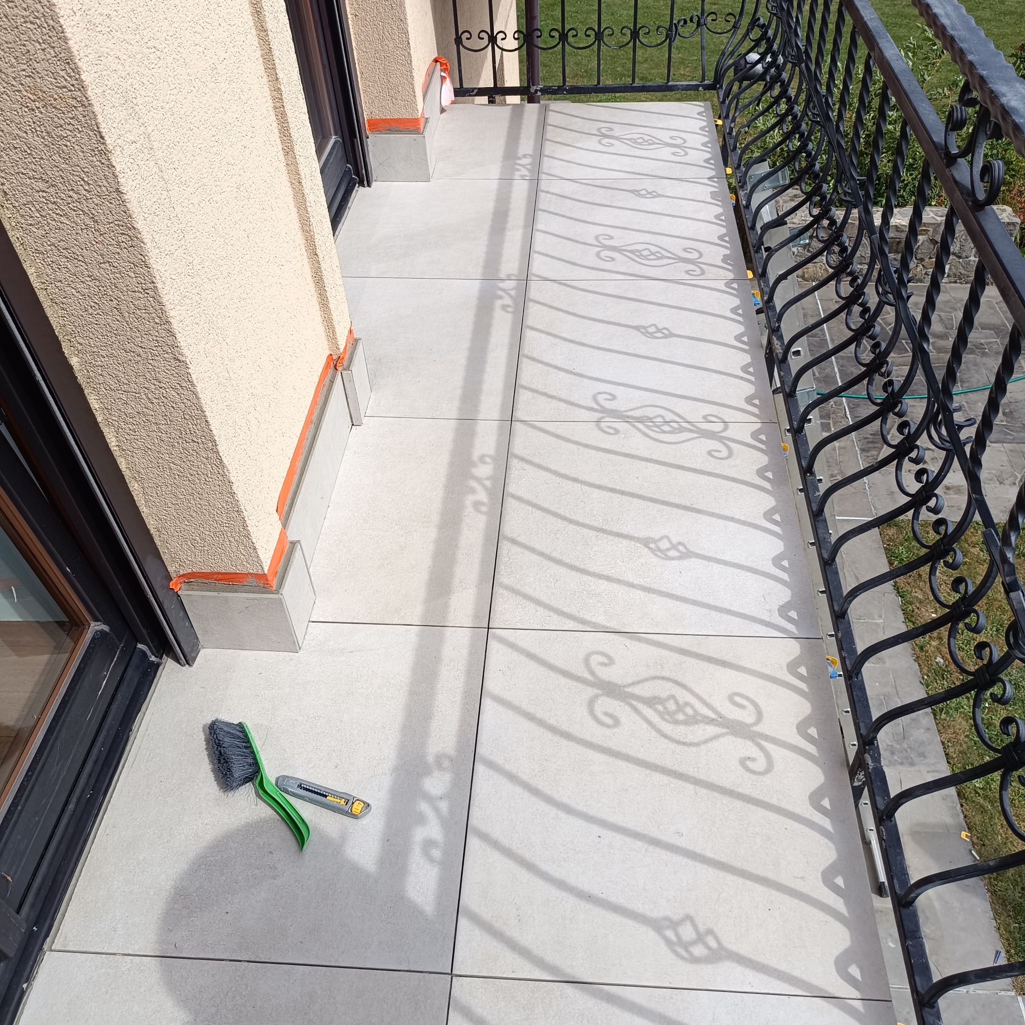 Wyłożony jasnymi płytkami balkon z ozdobną, kutą balustradą. Na podłodze leżą szczotka i nóż do cięcia. Widoczne cienie balustrady na płytkach.