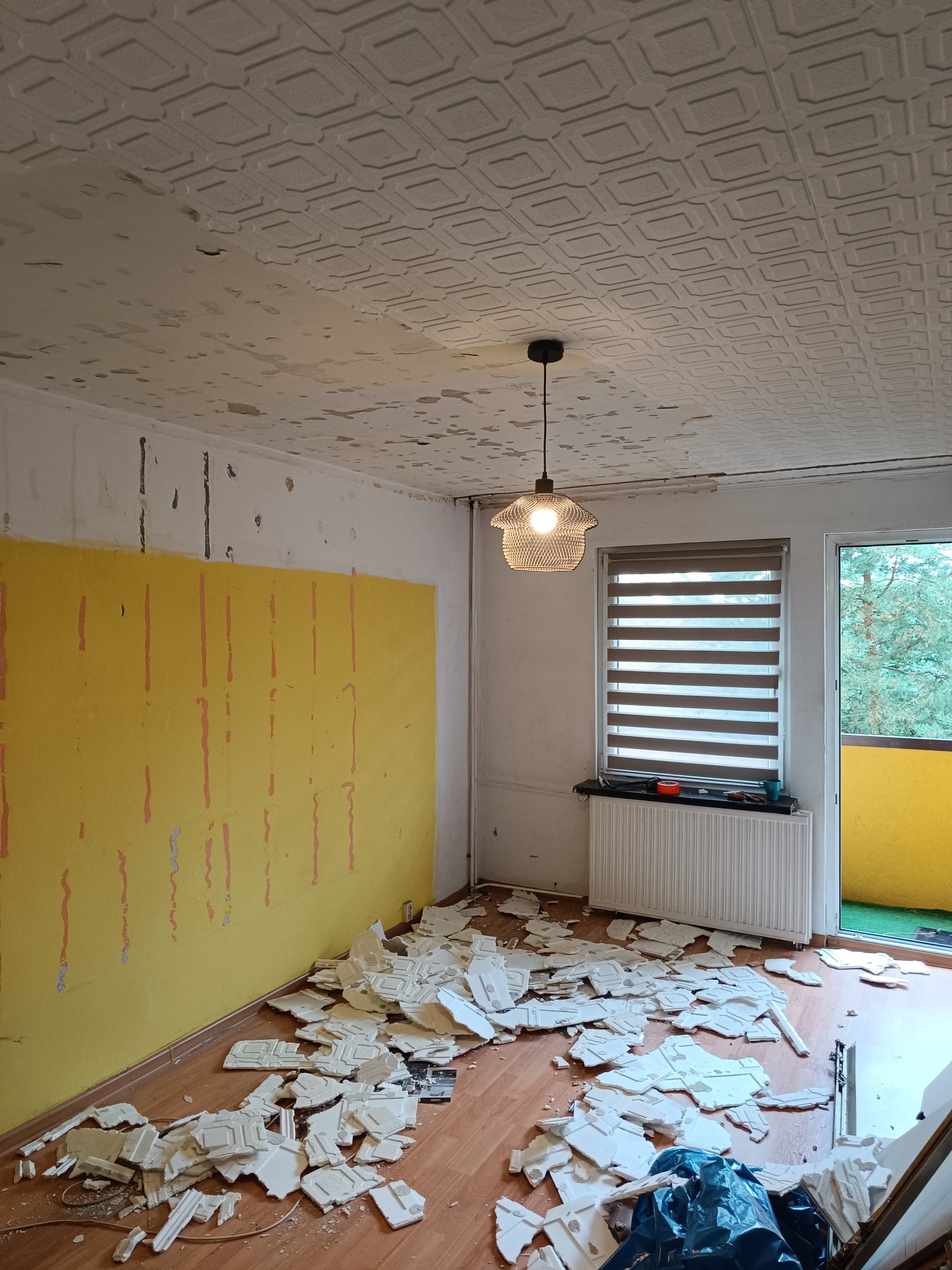 Pokój w trakcie demontażu sufitu kasetonowego. Widoczne fragmenty kasetonów na podłodze, odsłonięta żółta ściana i lampa wisząca. Balkon z zieloną podłogą w tle.