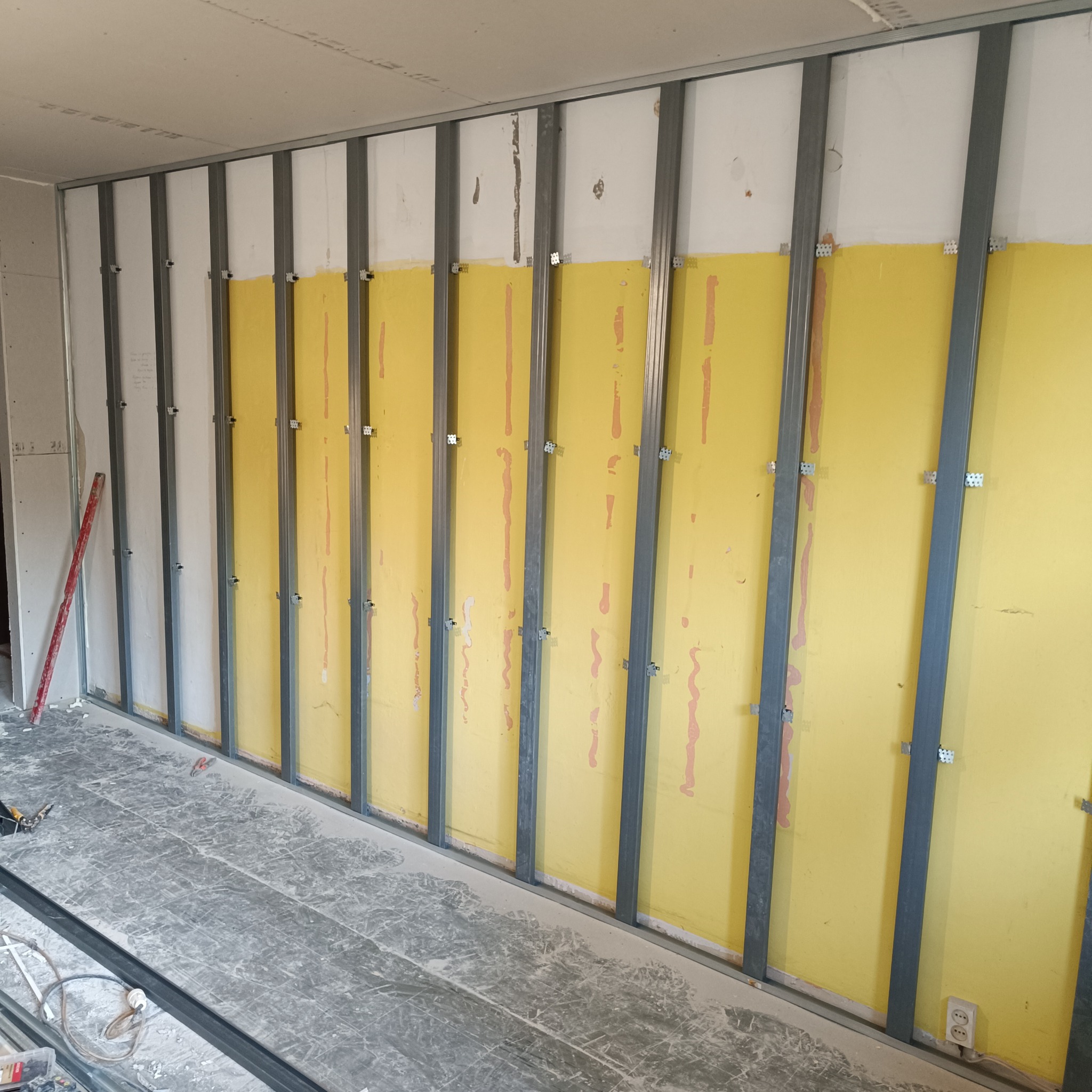 Montaż profili pod płyty gipsowo-kartonowe na ścianie z żółtą izolacją. Widoczne narzędzia i fragment podłogi w trakcie prac wykończeniowych.