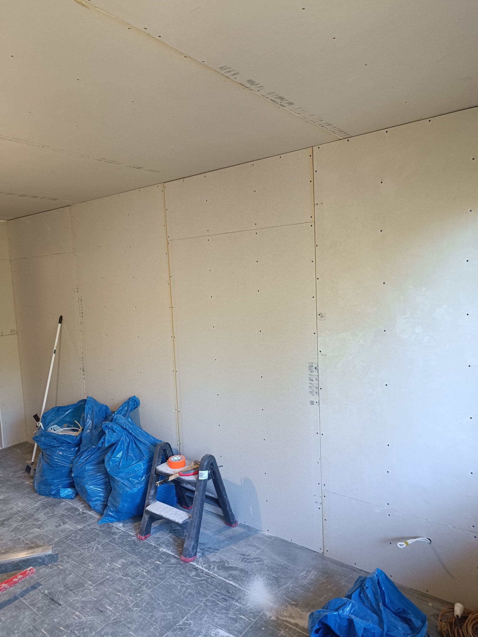 Montaż płyt karton-gips na ścianach i suficie. Widoczne narzędzia, drabina i worki z odpadami. Etap prac wykończeniowych w pomieszczeniu.