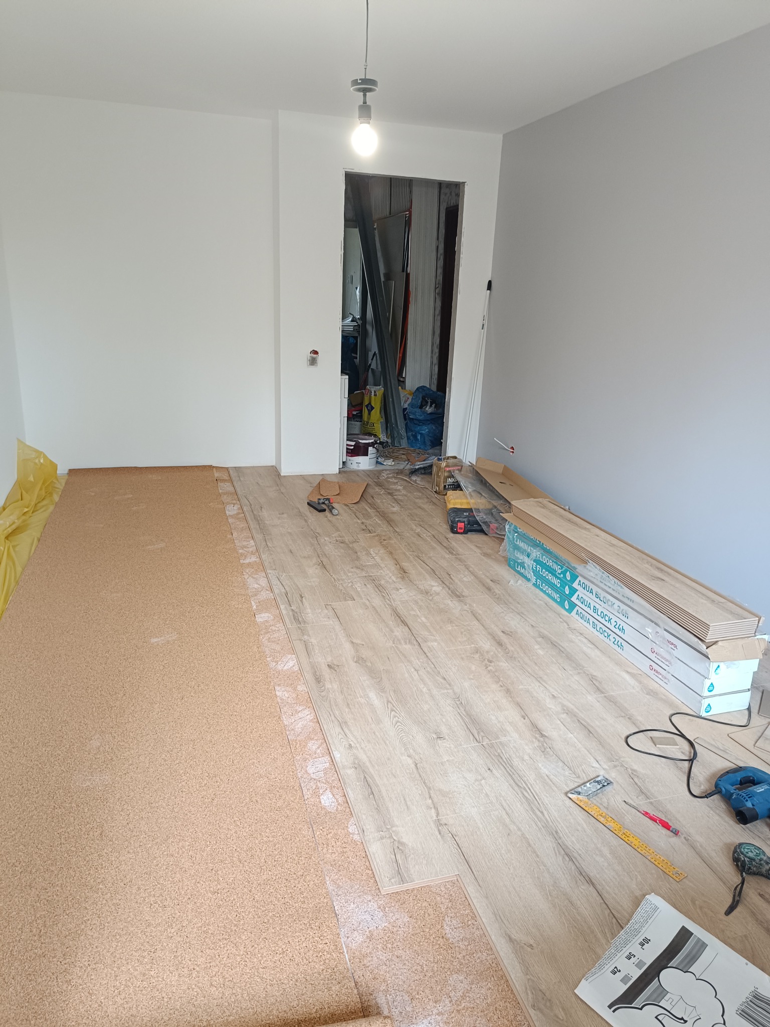Układanie paneli podłogowych w jasnym wnętrzu. Część podłogi wyłożona korkiem, obok narzędzia i paczki paneli Aqua Block 24h. Widoczne białe ściany i sufit.