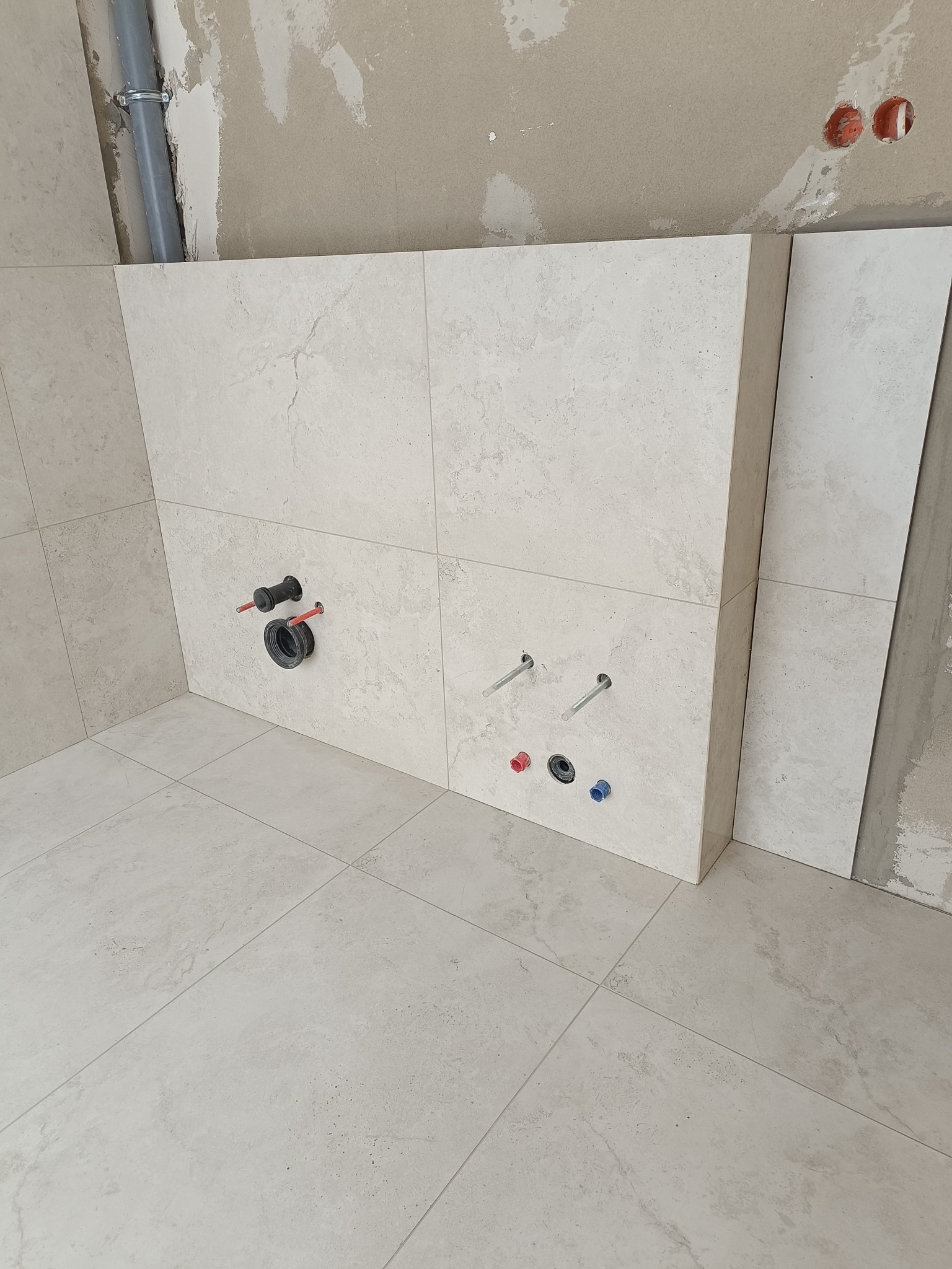 Wykończona ściana i podłoga w łazience z jasnych, kwadratowych płytek. Widoczne przyłącza wodne i kanalizacyjne. Ściana częściowo wykończona, surowy tynk w tle.