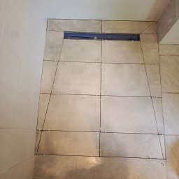 Tomasz Pietrzak - Ułożone płytki podłogowe w łazience z odpływem liniowym, widoczne krzyżyki dystansowe. Jasne płytki na ścianach i podłodze. Ukończony etap prac.