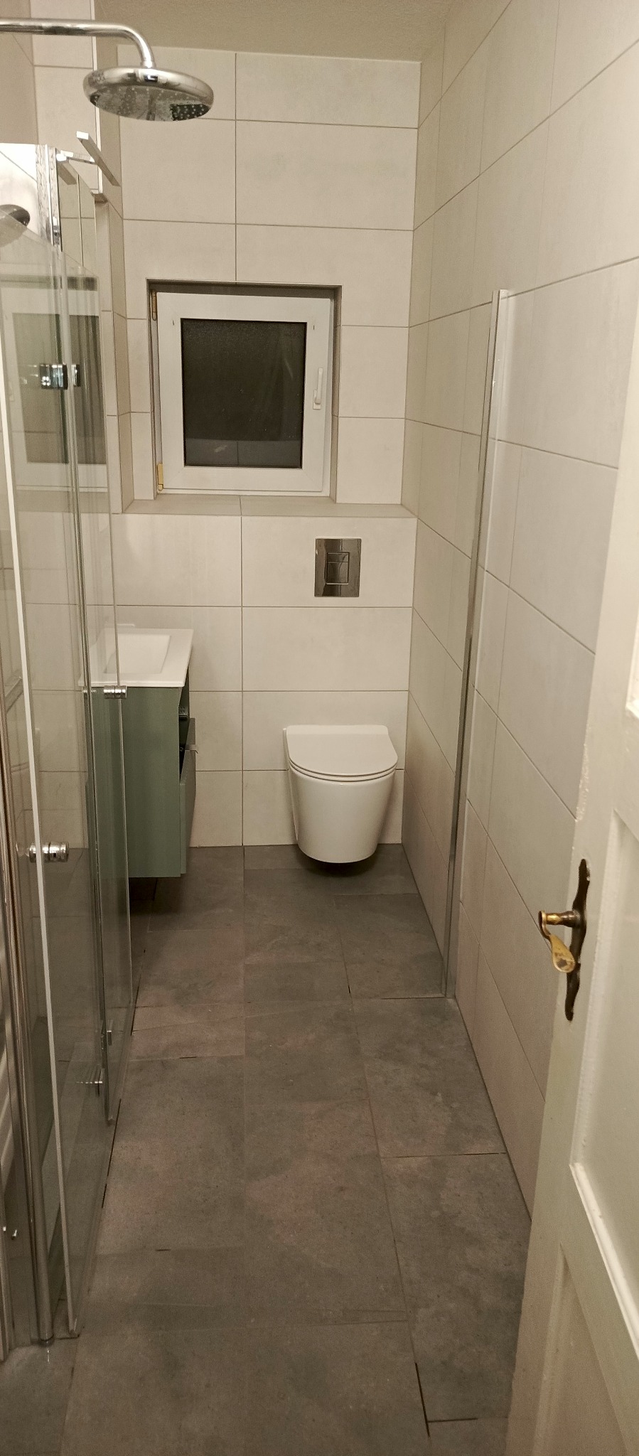 Jasna łazienka po remoncie: kabina prysznicowa ze szklanymi drzwiami, umywalka z szafką, podwieszana toaleta, okno i szare płytki podłogowe.