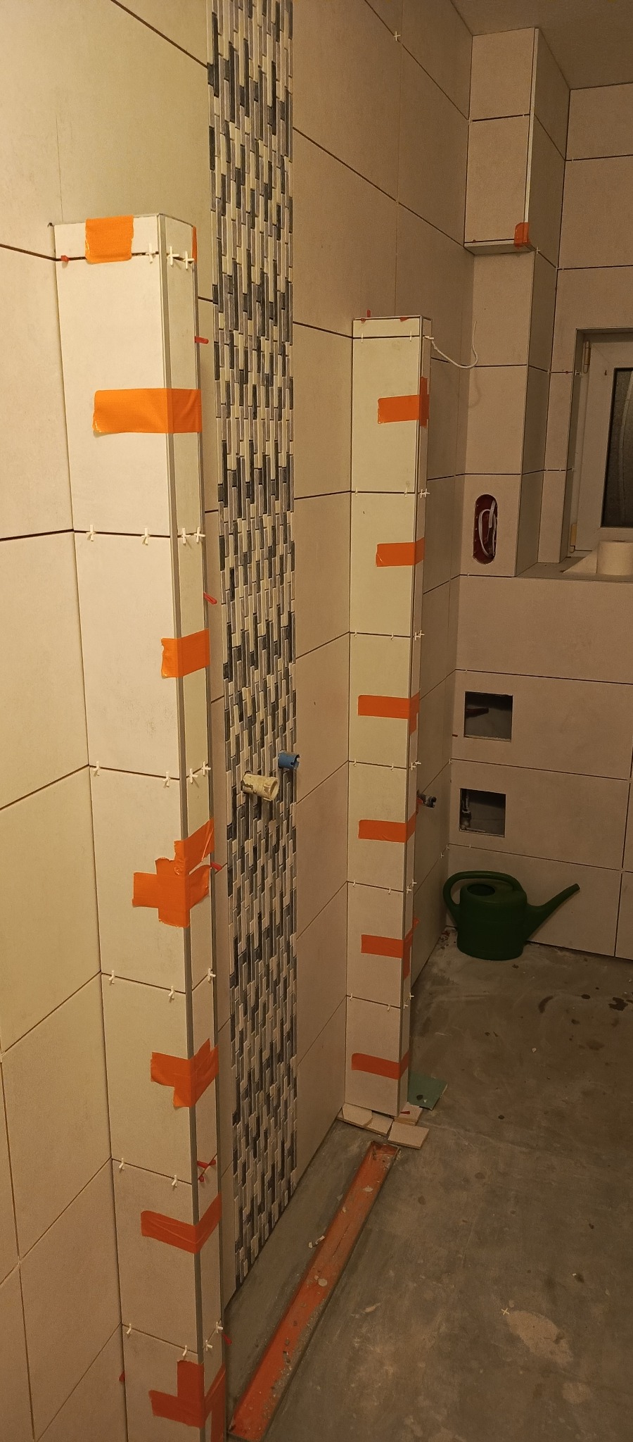 Wnętrze łazienki w trakcie prac: ułożone płytki ceramiczne na ścianach, zabezpieczone pomarańczową taśmą, widoczna mozaika dekoracyjna i poziomica.