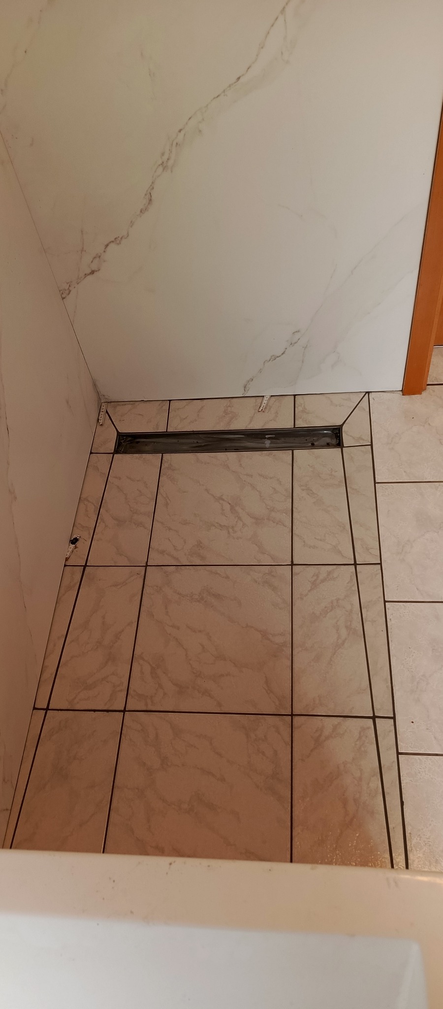 Ułożone płytki podłogowe w łazience z odpływem liniowym. Płytki imitujące marmur, jasne, z ciemnymi fugami. Widok z góry na fragment podłogi i ściany.