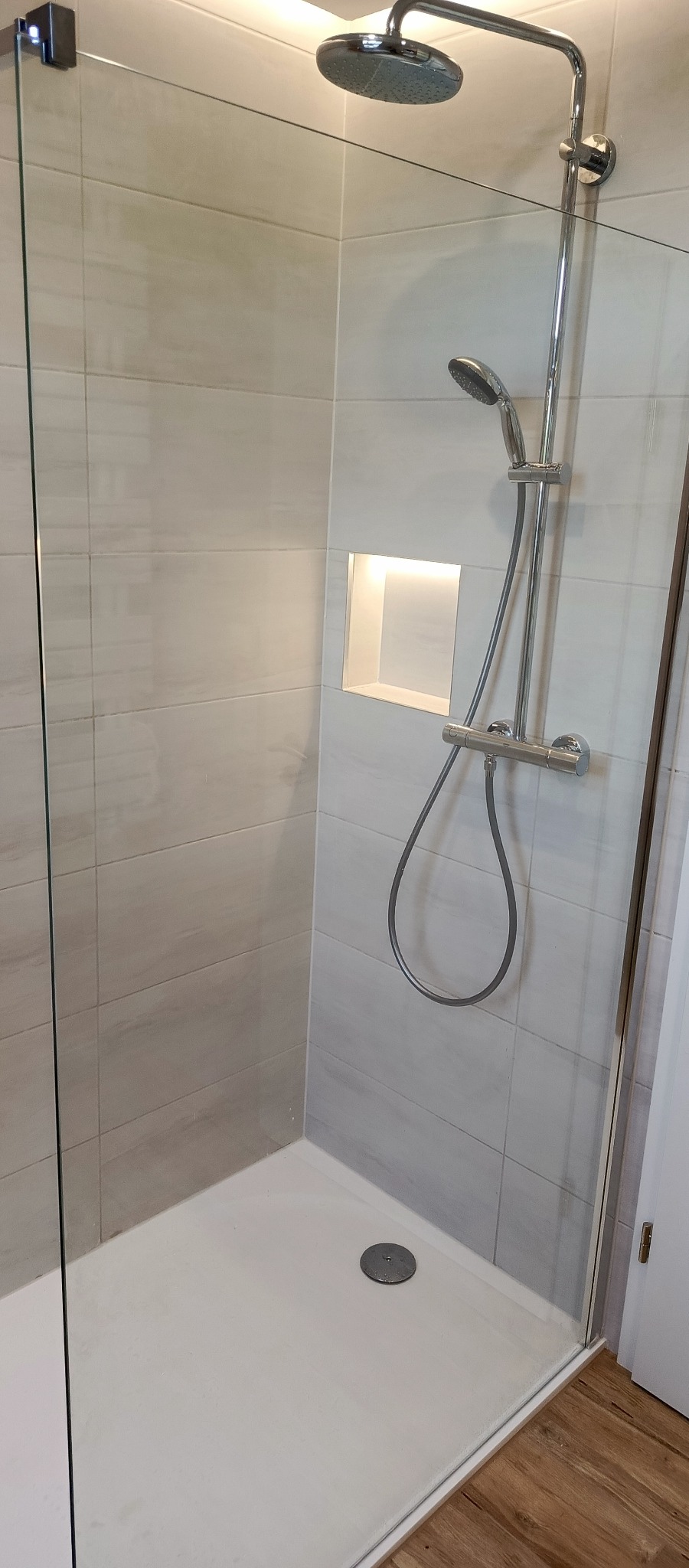 Nowoczesna kabina prysznicowa z deszczownicą i wnęką z podświetleniem w jasnej łazience. Widoczne szklane drzwi i drewnopodobna podłoga.