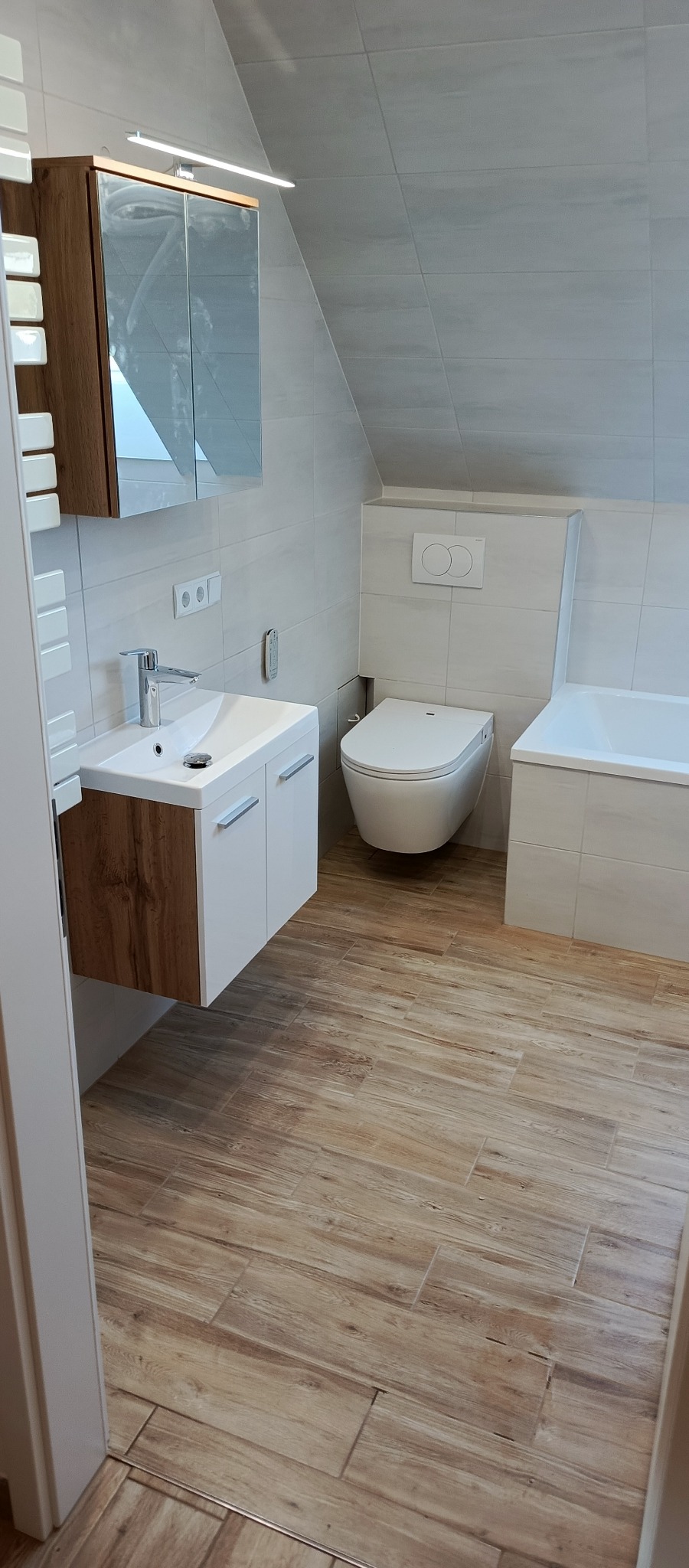 Jasna łazienka na poddaszu z drewnopodobną podłogą, umywalką z szafką i lustrem z oświetleniem. Uwagę przykuwa skośny sufit wykończony płytkami.