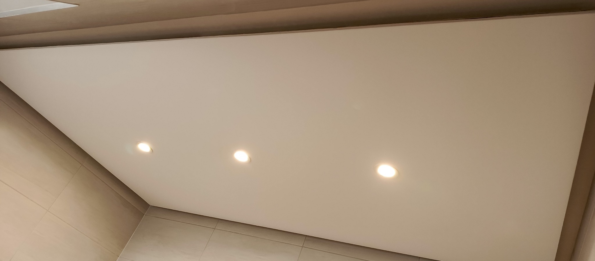 Podwieszany sufit w łazience z trzema punktowymi źródłami światła, beżowe płytki na ścianach, neutralna kolorystyka, widok z dołu.