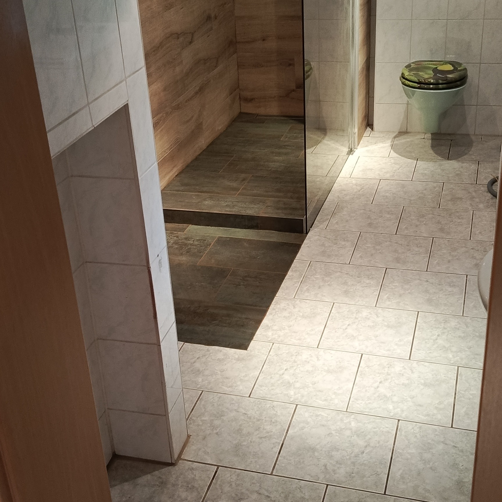 Wykończona łazienka z kabiną prysznicową typu walk-in, wyłożona płytkami w odcieniach szarości i brązu. Widoczna toaleta z oryginalną deską sedesową.