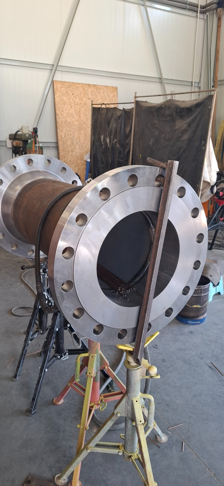 Spawanie rury z kołnierzami na stojakach w warsztacie. Widoczne elektrody spawalnicze wewnątrz rury. Metalowa konstrukcja w tle.