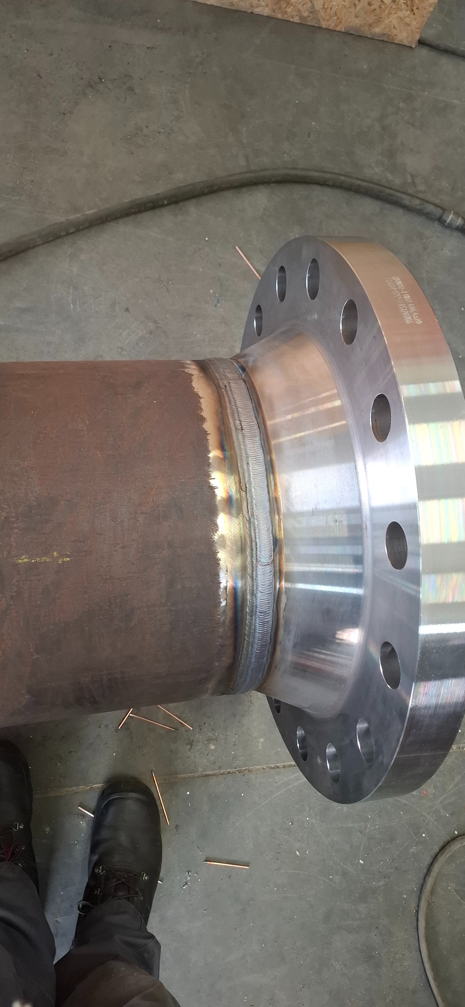 Spoina łącząca rurę z kołnierzem, widoczne ślady spawania i elektrody na podłodze. Buty robocze w dolnej części kadru. Metalowa konstrukcja.