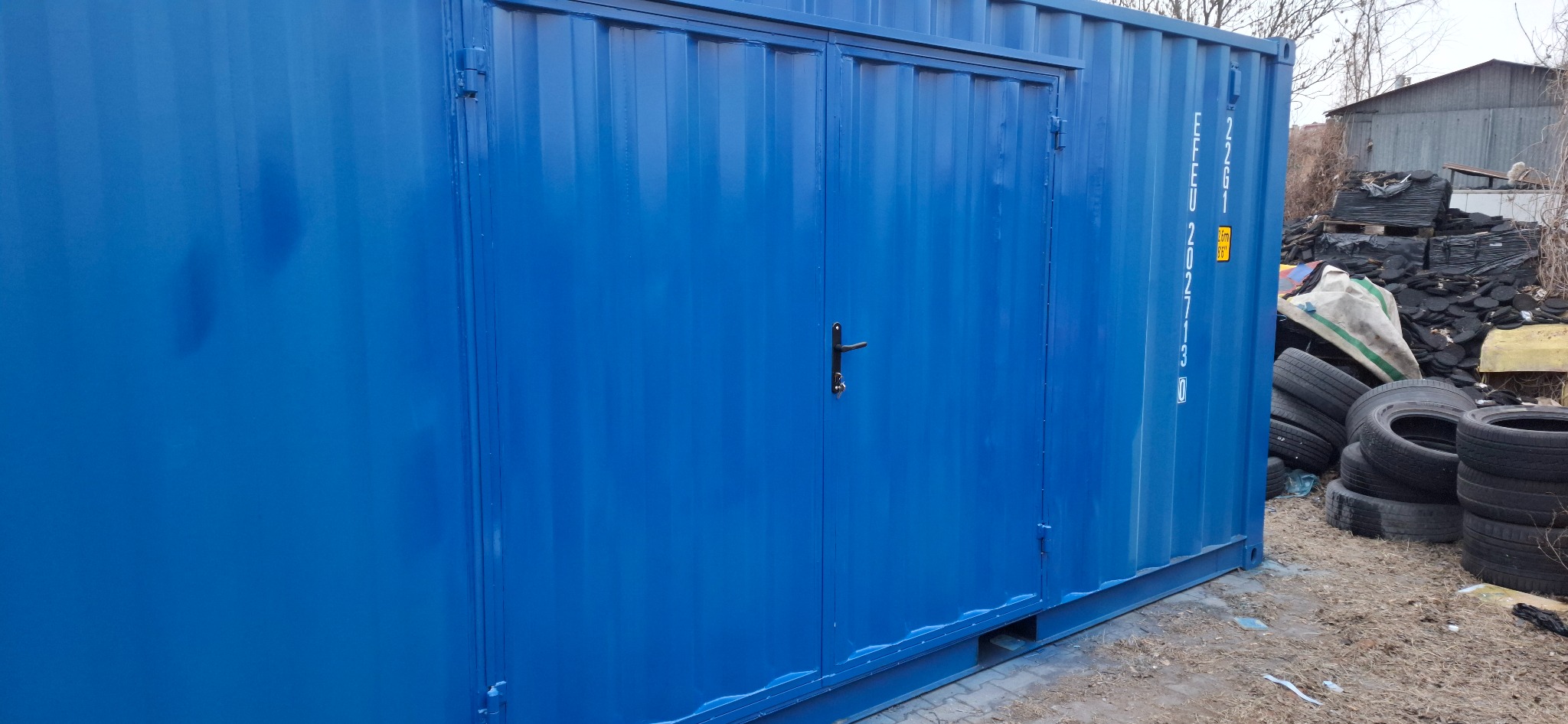 Niebieski kontener z metalowymi drzwiami i czarną klamką, widoczny z boku, z numerem seryjnym na ścianie. W tle składowisko opon i gruzu.