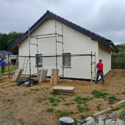 HANDEL OBWOŹNY - BOŻENA LADACH - Malowanie elewacji domu szkieletowego z użyciem rusztowań. Dwóch pracowników w trakcie pracy, widoczne materiały budowlane i narzędzia. Pochmurne niebo w tle.