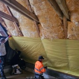 HANDEL OBWOŹNY - BOŻENA LADACH - Ocieplanie poddasza wełną mineralną i folią paroizolacyjną. Trzech mężczyzn w trakcie montażu, widoczne drewniane elementy konstrukcji dachu.