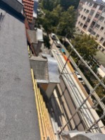 Montaż obróbek blacharskich na dachu budynku wielorodzinnego, widok z góry na ulicę i drzewa. Elementy rusztowania widoczne w kadrze.