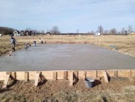 Zalana płyta fundamentowa pod dom szkieletowy, widoczne drewniane szalunki i wystające rury instalacyjne. W tle łąka i zabudowania.