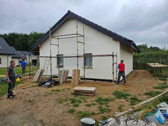 Malowanie elewacji domu szkieletowego z użyciem rusztowań. Dwóch pracowników w trakcie pracy, widoczne materiały budowlane i narzędzia. Pochmurne niebo w tle.