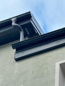 Szczegółowe ujęcie grafitowej rynny i okapu na tle jasnego nieba, widoczne mocowania. Element domu szkieletowego z bliska.