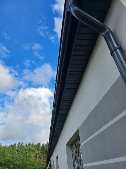 Szary dom z czarną rynną na tle błękitnego nieba z chmurami. Widok z dołu, podkreślający nowoczesny design i dbałość o detale wykończenia.