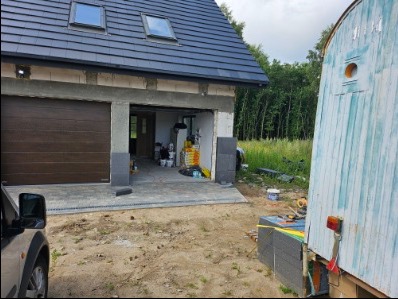 Nowoczesny dom w trakcie budowy z dwoma bramami garażowymi, jedna zamknięta brązowa, druga otwarta ukazująca wnętrze. Widoczne materiały budowlane i przyczepa z boku.