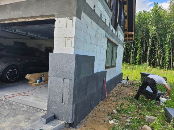 Ocieplanie ścian zewnętrznych domu z betonu komórkowego szarym styropianem grafitowym. Prace wykończeniowe na budowie, widoczny pracownik i garaż z samochodem.