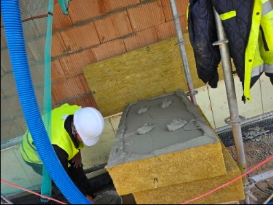 Pracownik w kasku i kamizelce montuje izolację z wełny mineralnej z zaprawą na ścianie z cegły, widoczne rusztowanie i niebieski przewód wentylacyjny.