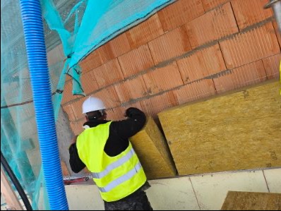 Pracownik w kasku i kamizelce montuje izolację z wełny mineralnej na ceglanej ścianie budynku w trakcie budowy. Widoczna niebieska rura wentylacyjna.