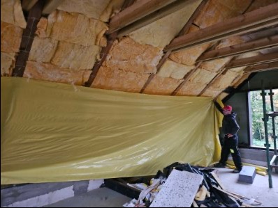 Ocieplenie poddasza wełną mineralną w domu szkieletowym, zabezpieczone folią. Widoczny fragment okna i pracownik w stroju roboczym.