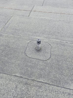 Szary dach pokryty papą z zamontowanym odpowietrznikiem w metalowej obudowie. Widoczne łączenia papy. Ujęcie z góry, bliski plan.