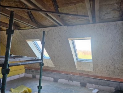 Płyta OSB na poddaszu z oknami dachowymi, widok z rusztowania. Izolacja stropu widoczna. Etap wykończenia poddasza w domu szkieletowym.