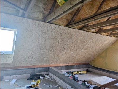 Skośna ściana poddasza wykończona płytą OSB, widoczne elementy konstrukcji dachu i okno połaciowe. Surowy stan, przygotowanie do dalszych prac wykończeniowych.