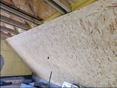 Wnętrze poddasza w trakcie wykańczania: płyta OSB na skośnej ścianie, widoczne elementy konstrukcji dachu i izolacja. Surowy stan, przygotowanie do dalszych prac.