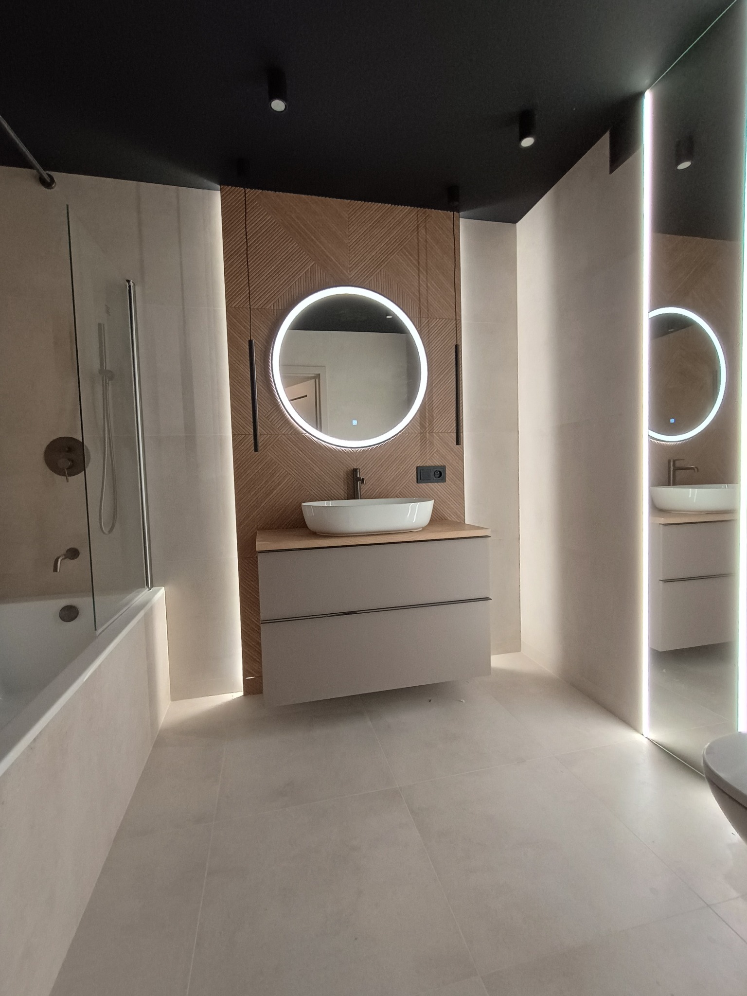 Nowoczesna łazienka z okrągłym lustrem LED, drewnianą ścianą i beżową szafką pod umywalką. Minimalistyczny design z oświetleniem punktowym i jasnymi płytkami.