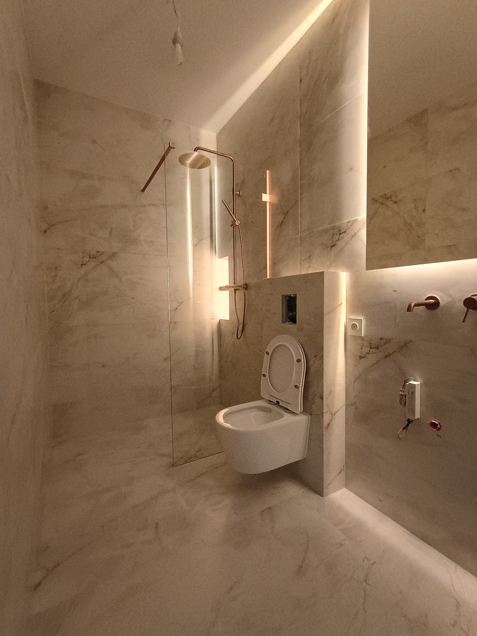 Wykończona łazienka z marmurowymi płytkami, podświetleniem LED i złotą armaturą. Widoczna toaleta podwieszana, kabina prysznicowa i elementy instalacji.