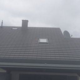 Michał - Szary dach z dachówką ceramiczną, komin, okna dachowe i antena satelitarna na tle pochmurnego nieba. Widok z dołu, fragment budynku w dolnej części kadru.