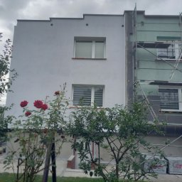 Michał - Dom w trakcie ocieplania: jedna ściana pomalowana na biało, druga w trakcie ocieplania styropianem, widoczne rusztowania. W tle pochmurne niebo i roślinność.