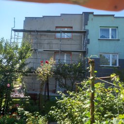 Michał - Budynek w trakcie prac elewacyjnych, widoczne rusztowania i fragment ogrodu z kwitnącymi różami. Część budynku otynkowana, druga pomalowana na zielono.