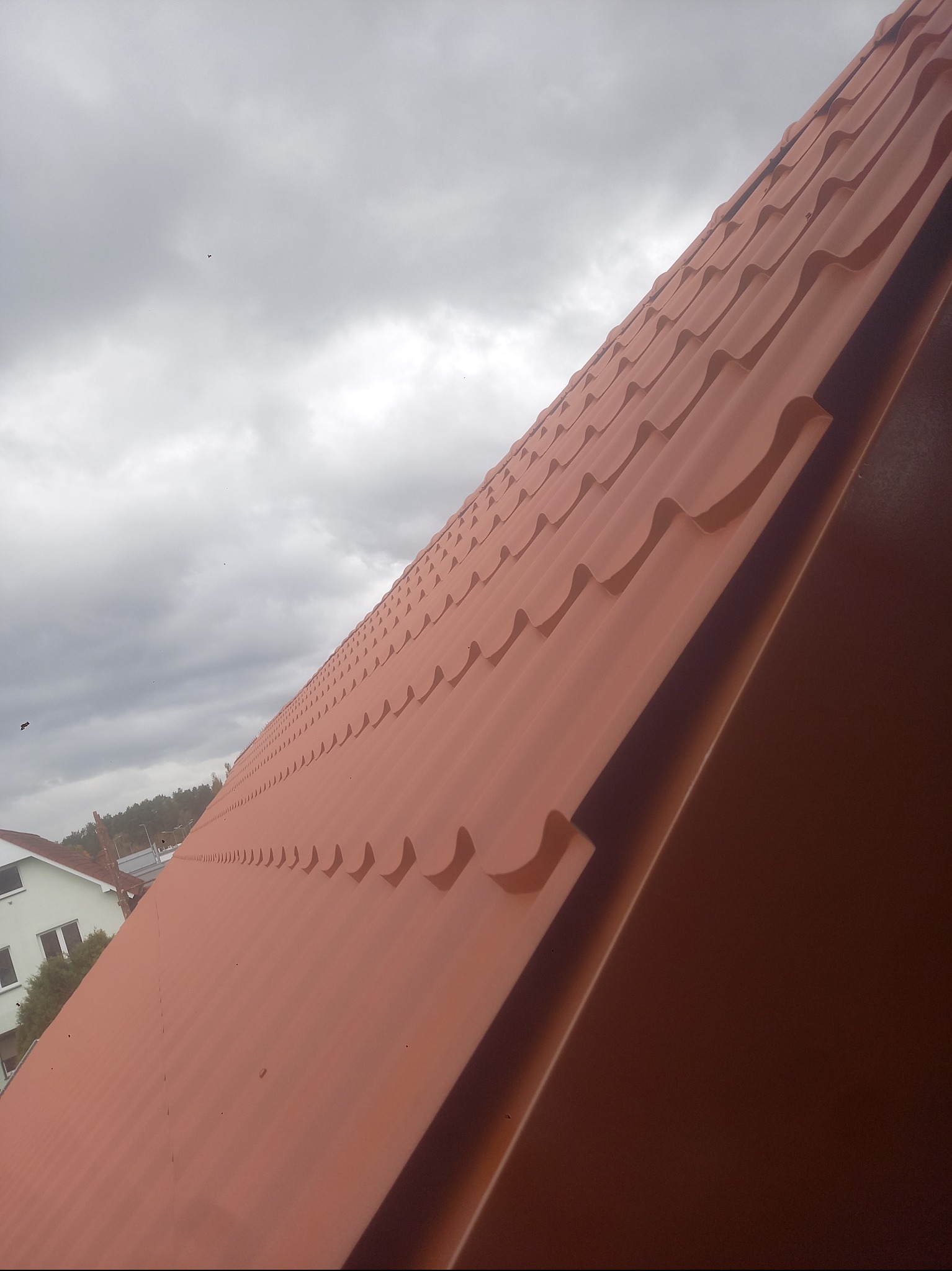 Czerwona blachodachówka na skośnym dachu domu, widziana pod kątem, z fragmentem budynku i pochmurnym niebem w tle. Widoczne regularne fale blachy.