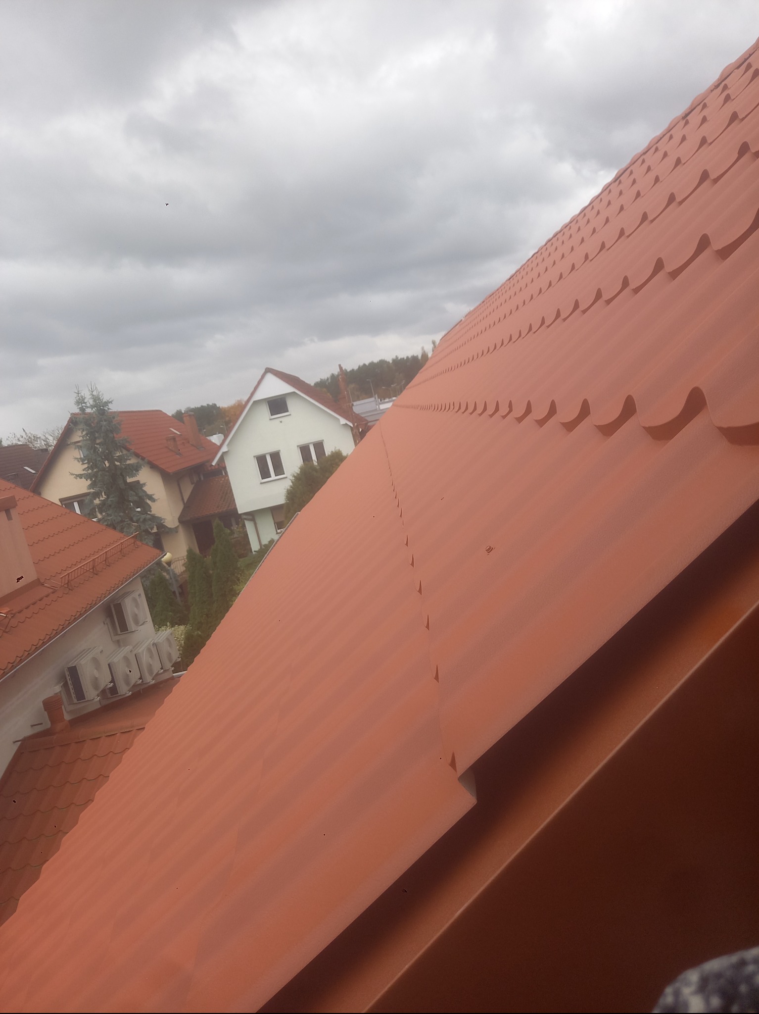 Czerwona blachodachówka na skośnym dachu domu jednorodzinnego, widok z góry, w tle zabudowa mieszkalna i pochmurne niebo. Charakterystyczny wzór blachy.