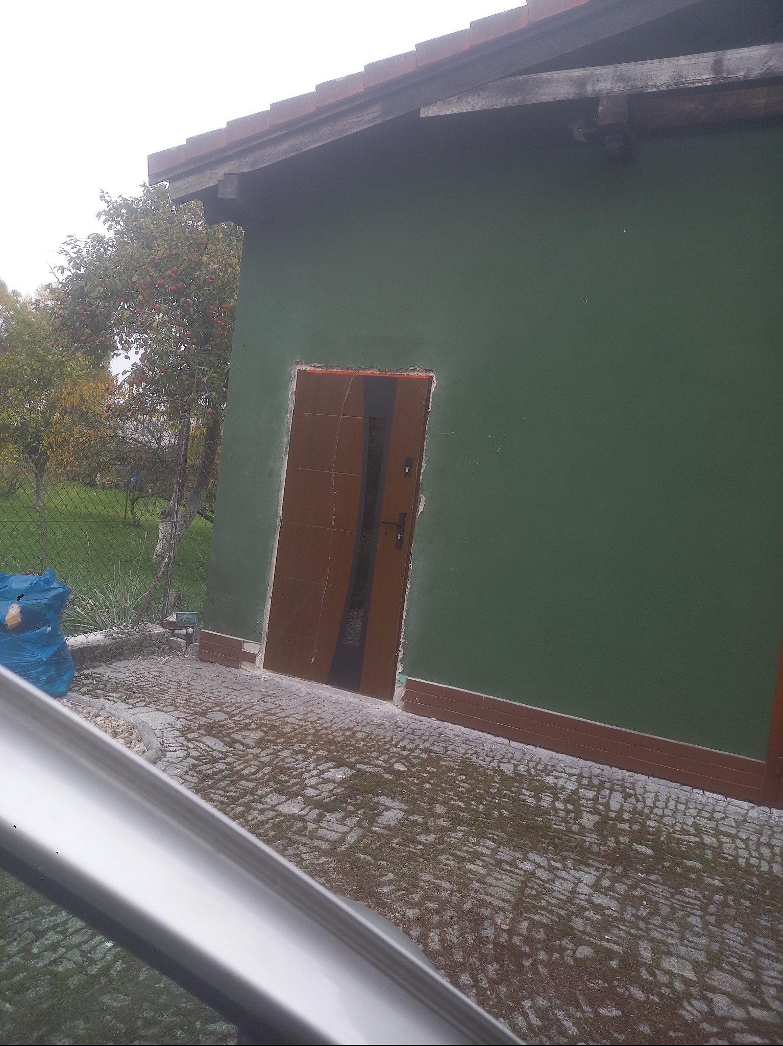 Zielona ściana budynku z brązowymi elementami wykończeniowymi i brązowymi drzwiami. Widoczny fragment chodnika z kostki brukowej i drzewo z czerwonymi owocami w tle.