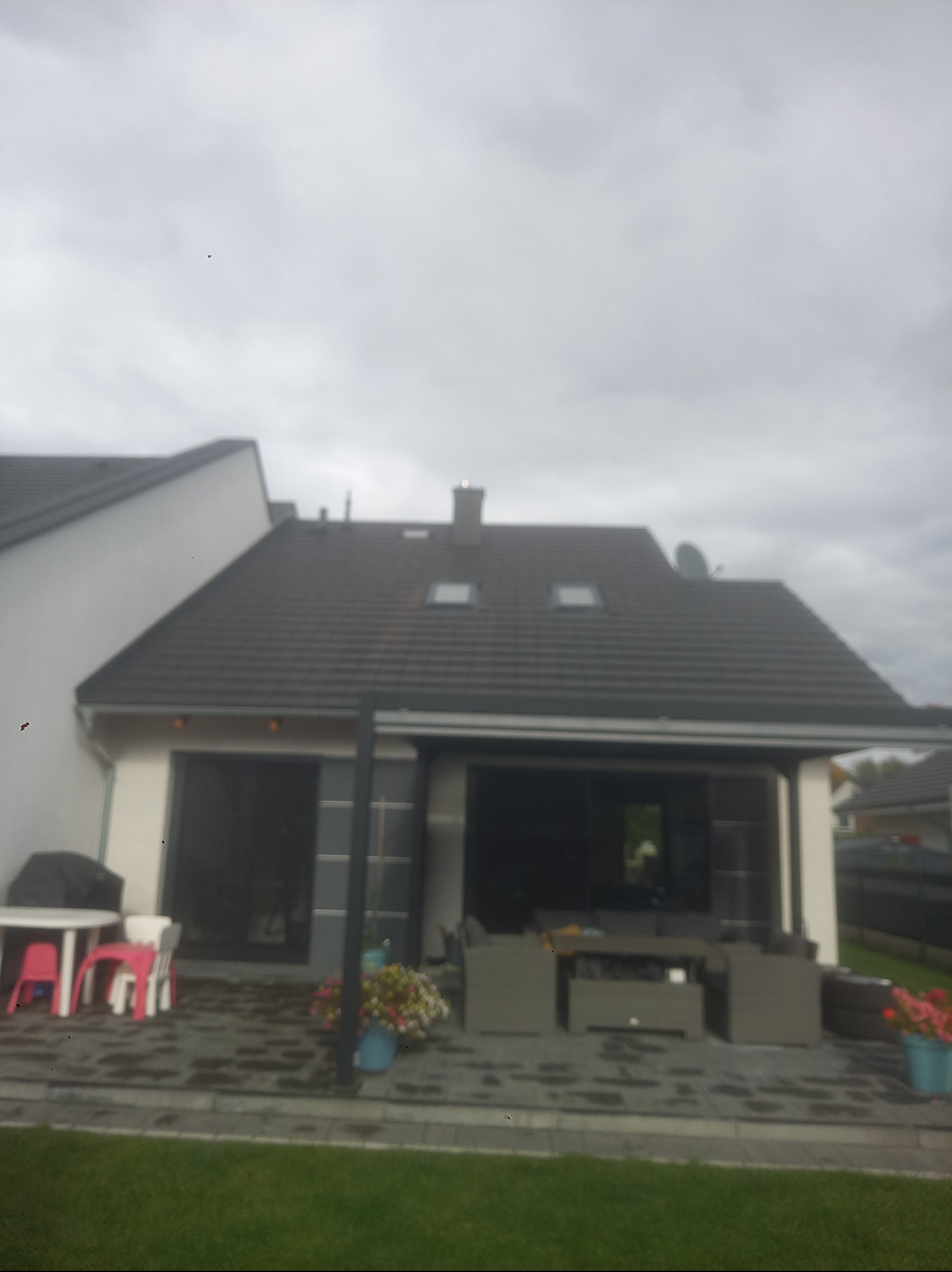 Nowoczesny dom jednorodzinny z ciemną dachówką i tarasem. Widoczne meble ogrodowe i kwiaty w donicach. Pochmurne niebo w tle.