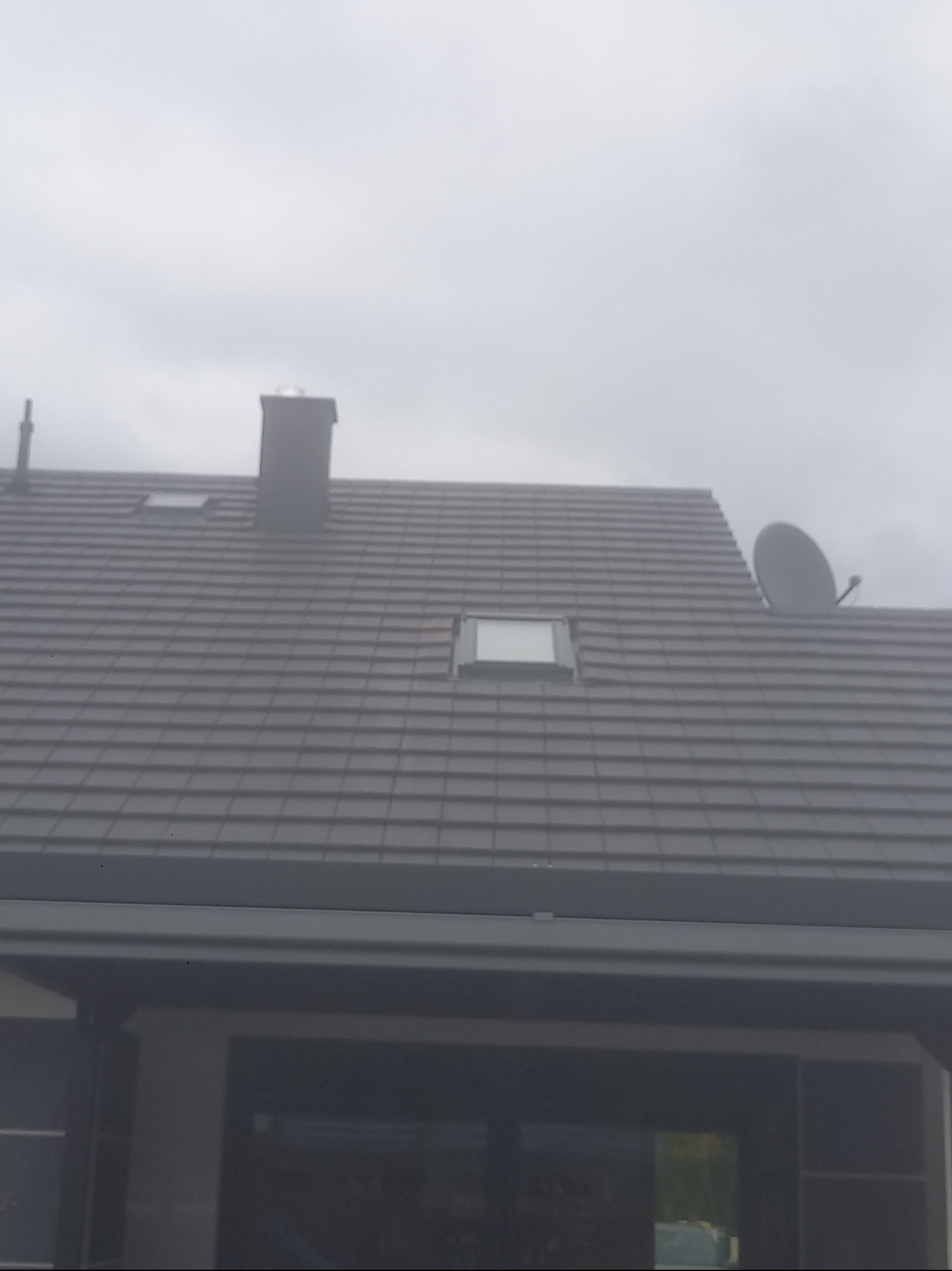 Szary dach z dachówką ceramiczną, komin, okna dachowe i antena satelitarna na tle pochmurnego nieba. Widok z dołu, fragment budynku w dolnej części kadru.