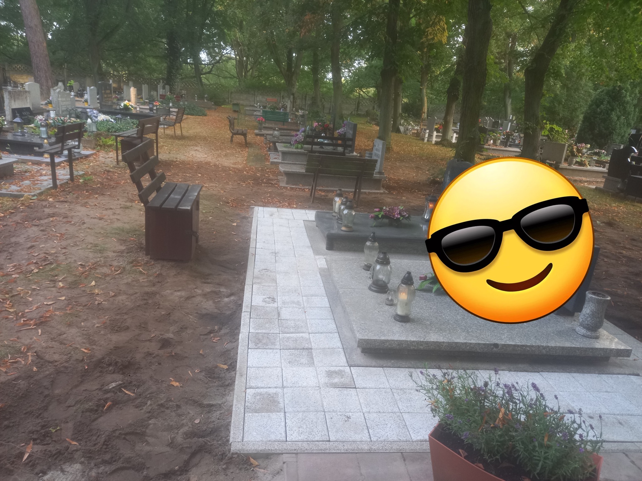 Ułożona kostka brukowa wokół nagrobka na cmentarzu, z widocznymi zniczami i kwiatami. Tło stanowią drzewa i inne nagrobki.